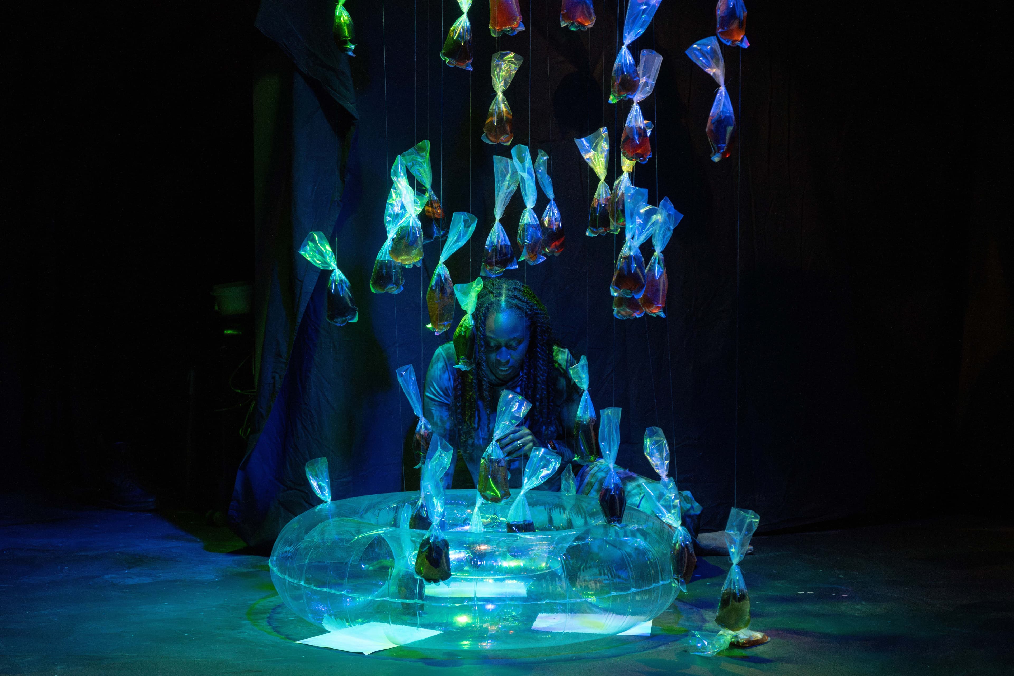 Maryam Kazeem is kneeling in front of a transparent inner tube. Plastic bags filled with water and trash are suspended above, reflecting the blue and green light.