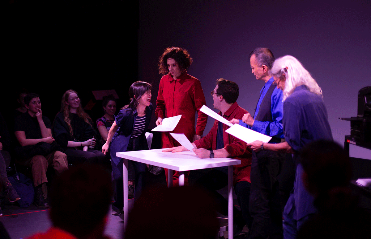 A woman in a stripped t-shirt yelling at a man in a red shirt sitting at a white table. A woman in a red jumpsuit looks over them. A man and woman wearing blue stand to the side. They are all holding sheets of paper.