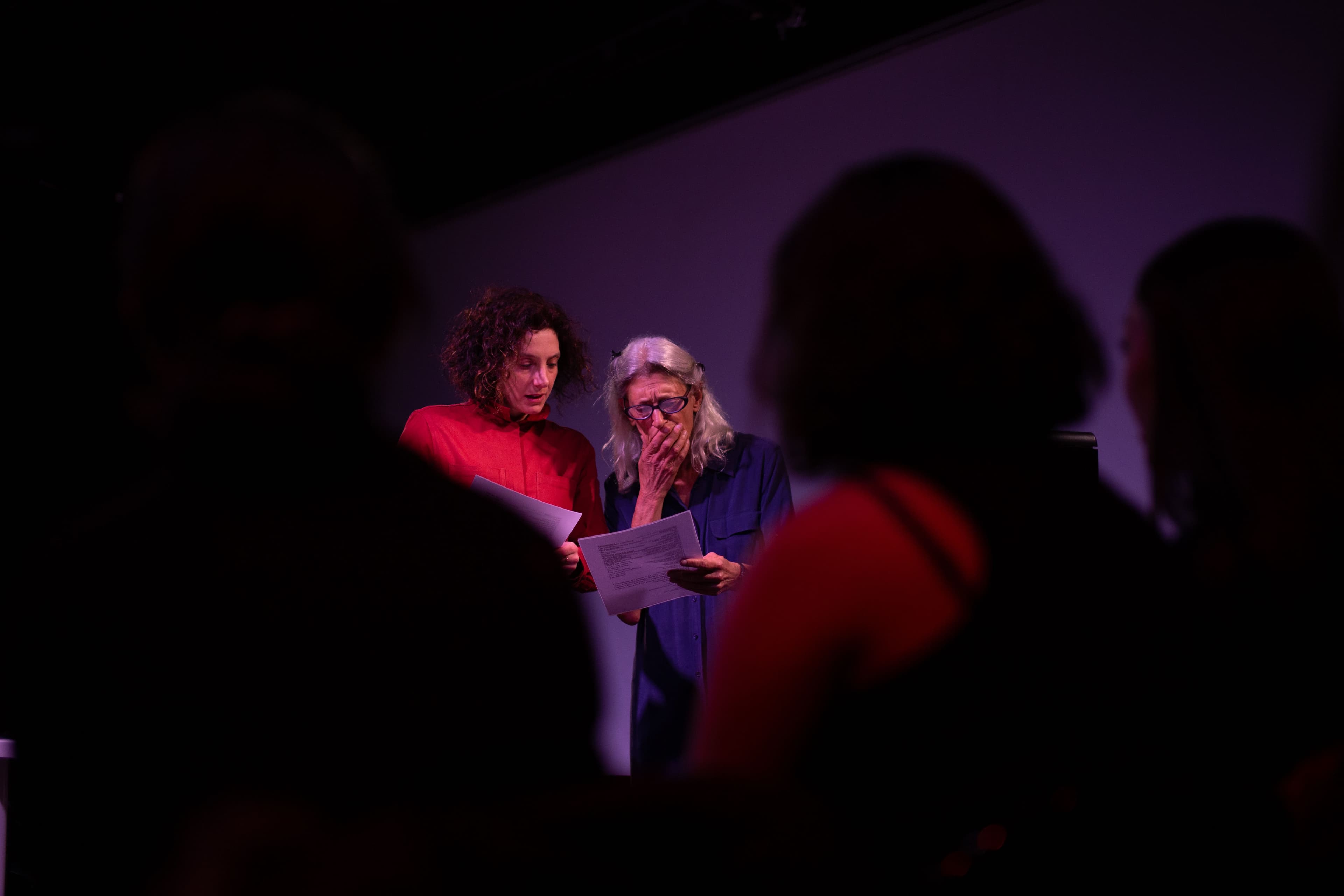 Framed in between the heads of an audience, focus on a woman wearing a red jumpsuit next to a woman with long grey hair in a blue jump suit covering her mouth. They are both looking at a sheet of paper.