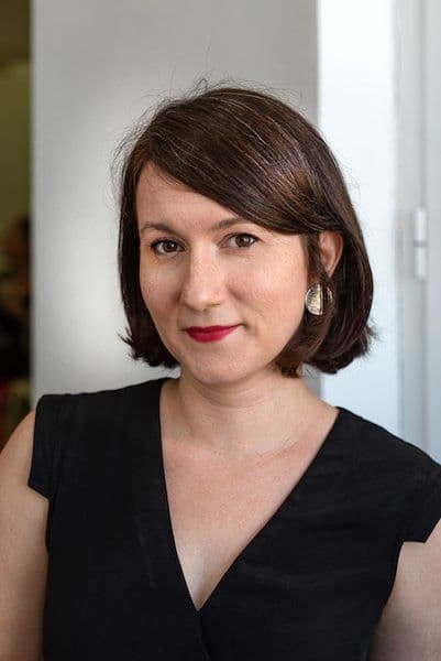 A white woman smiles at the camera, wearing black standing against a white backdrop.