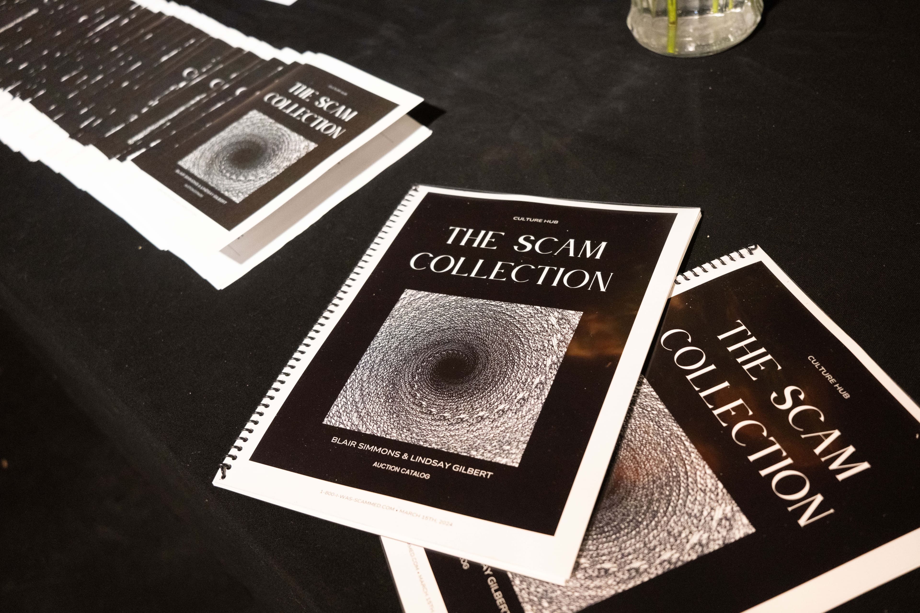 Two spiral bound copies of a catalogue titled The Scam Collection sit on a black table, next to a row of paper programs with the same cover.