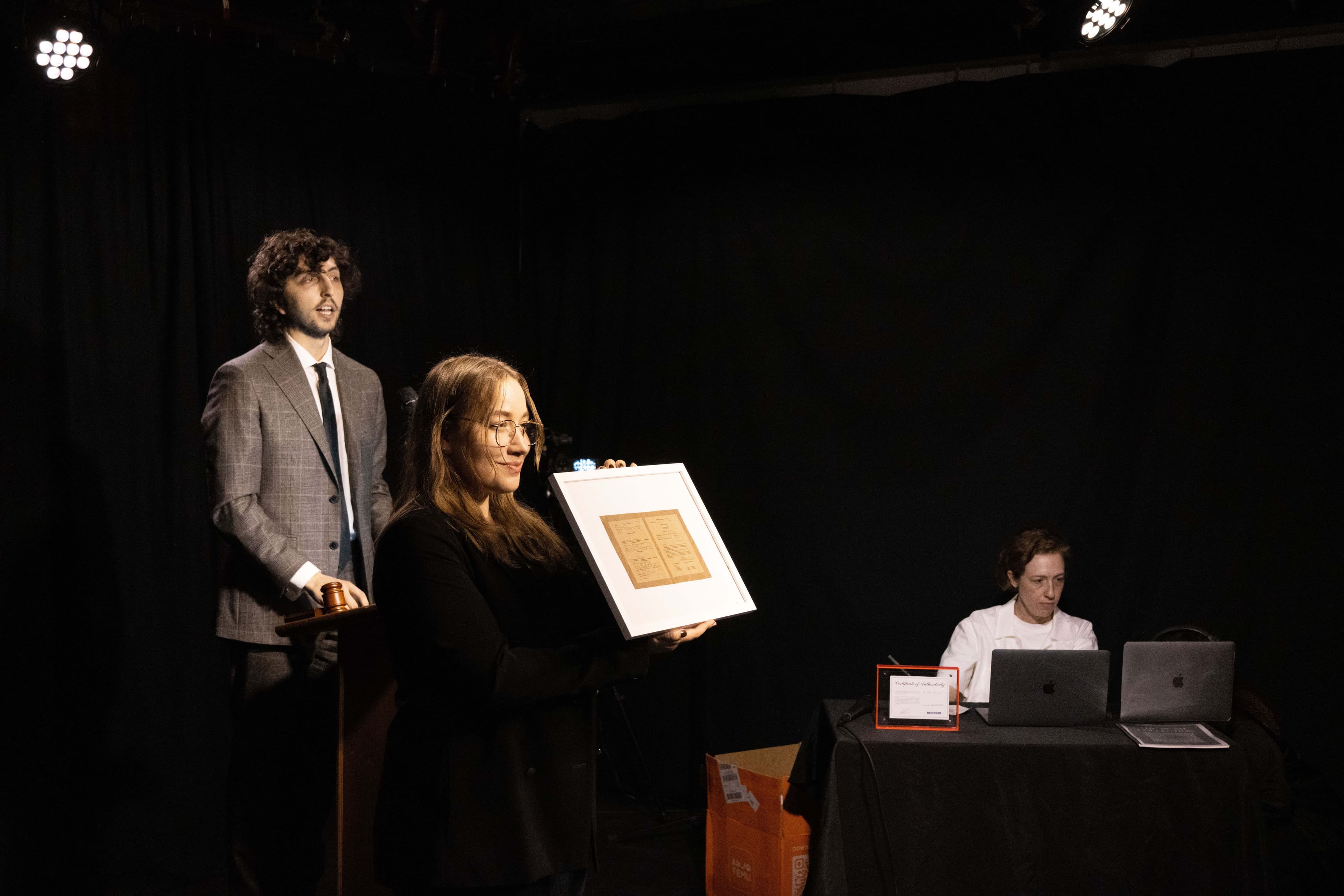 The artist Blair Simmons, a white woman with brown hair and glasses, holds up a framed print. Next to her, the auctioneer Hal Schulman wears a grey suit and tie. At the table, Lindsay Gilbert in a white t-shirt sits at a table with two laptops.