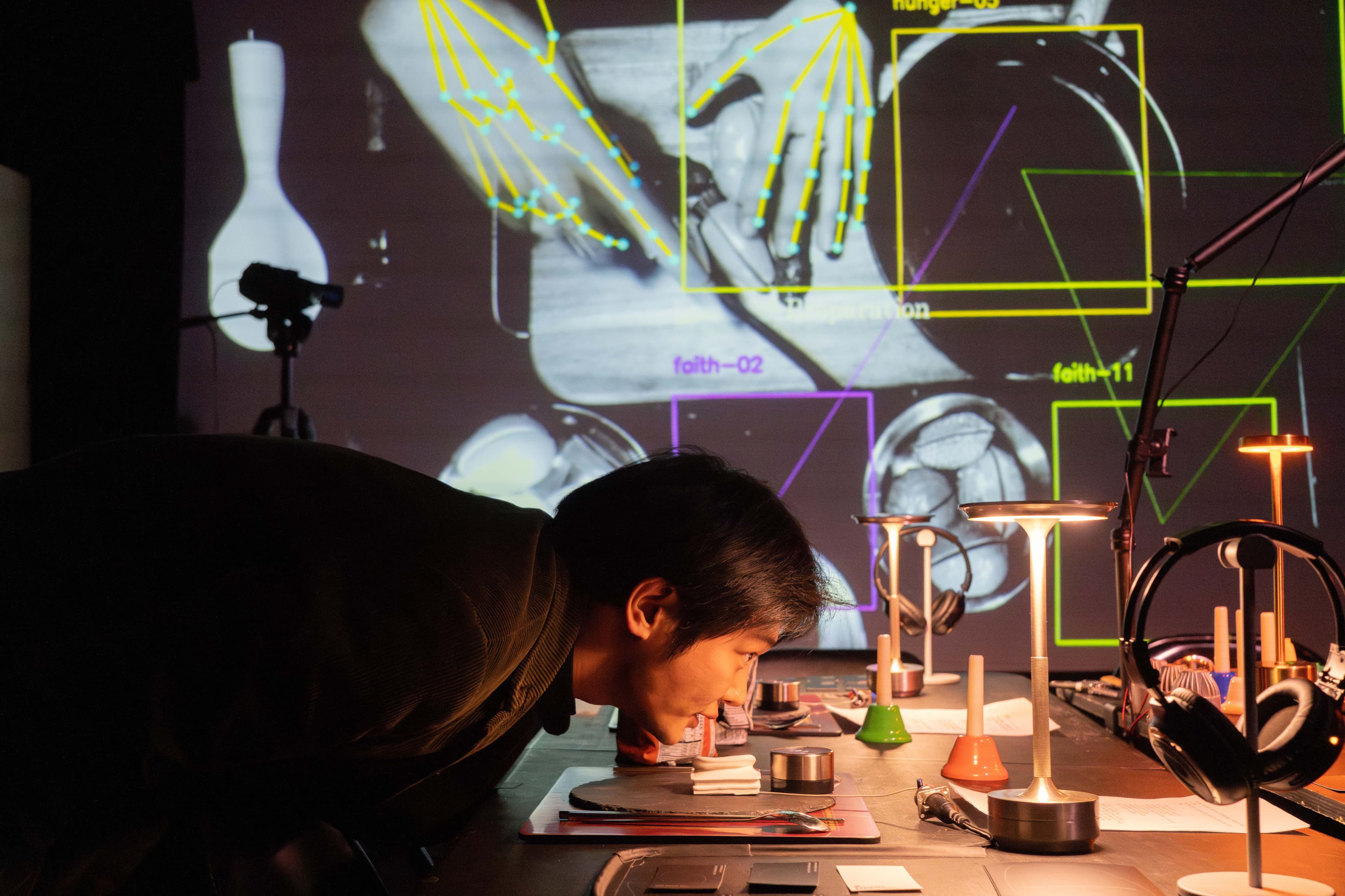 A man leaning over a table above a cardboard plate with a model ear on it. Behind him on the screen is a birdseye view of hands cutting fake fruit overlayed with machine vision lines.