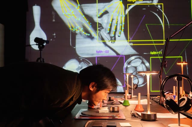 A man leaning over a table above a cardboard plate with a model ear on it. Behind him on the screen is a birdseye view of hands cutting fake fruit overlayed with machine vision lines.