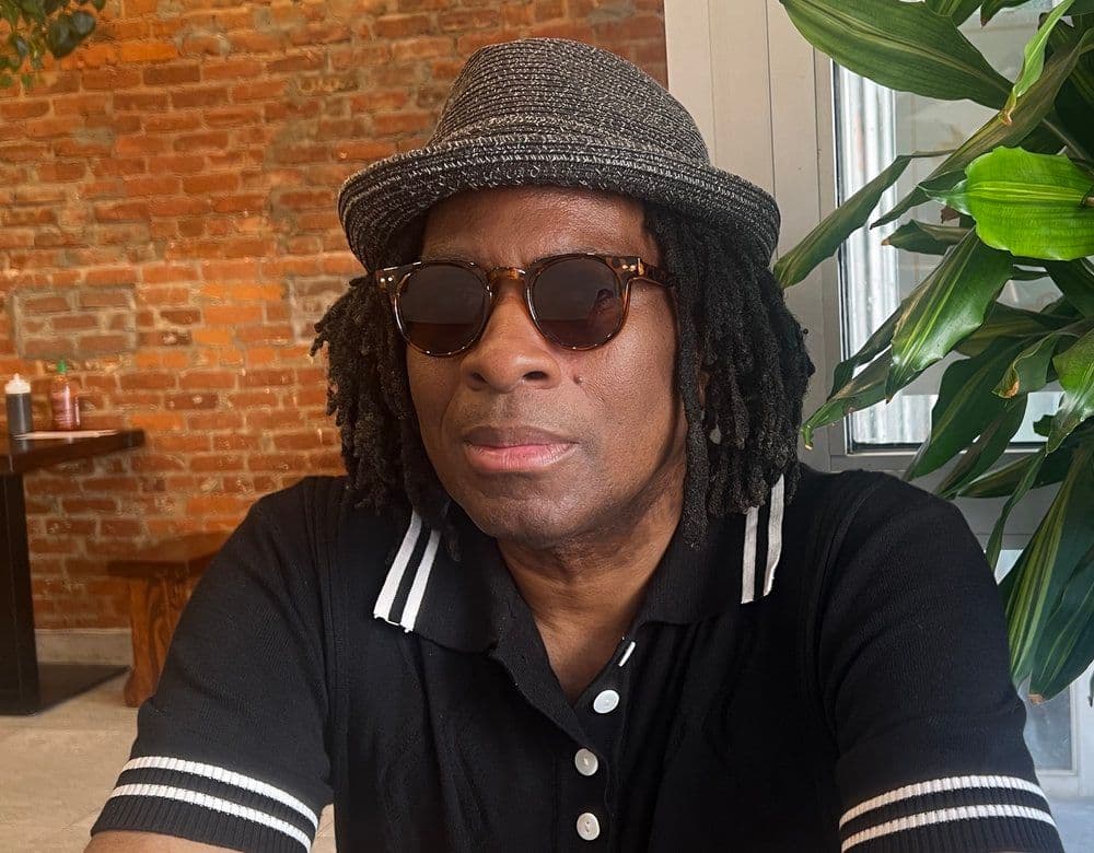 A Black man wearing sunglasses, a woven hat, and a striped polo shirt looks at the camera in front of a brick wall.