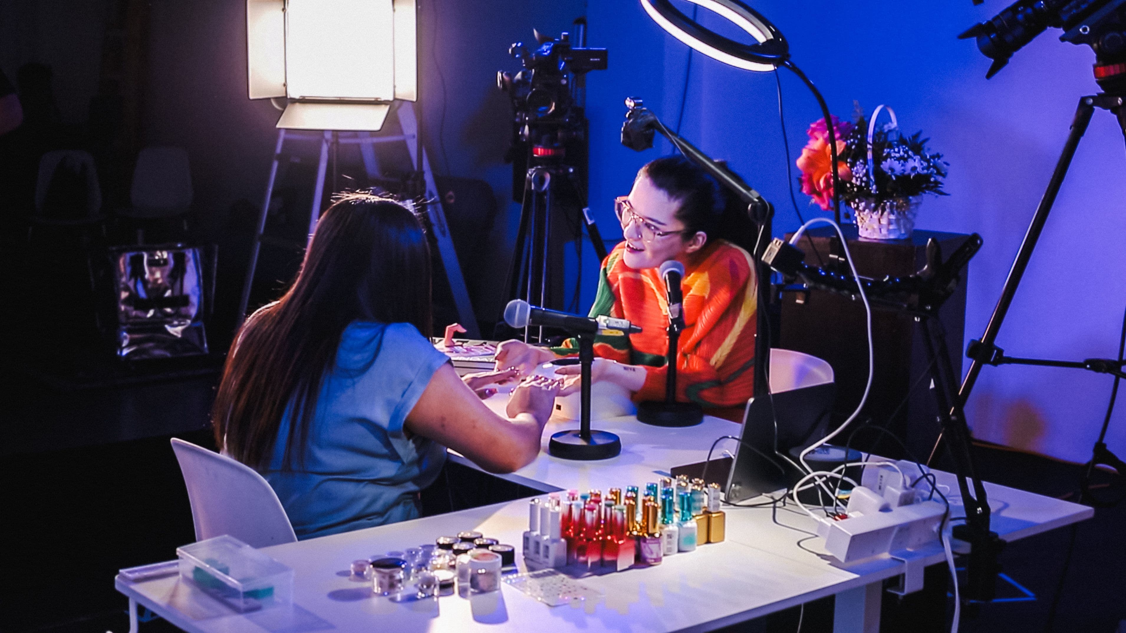 Arabelle Sicardi sits at a table across from a nail technician who is doing her nails. They are engaged in conversation. Microphones, filming equipment, and nail products surround them.