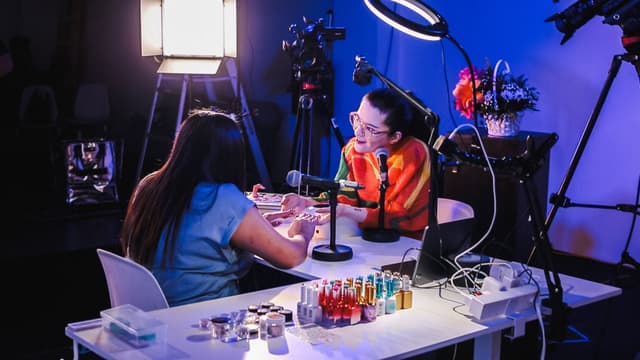 Arabelle Sicardi sits at a table across from a nail technician who is doing her nails. They are engaged in conversation. Microphones, filming equipment, and nail products surround them.