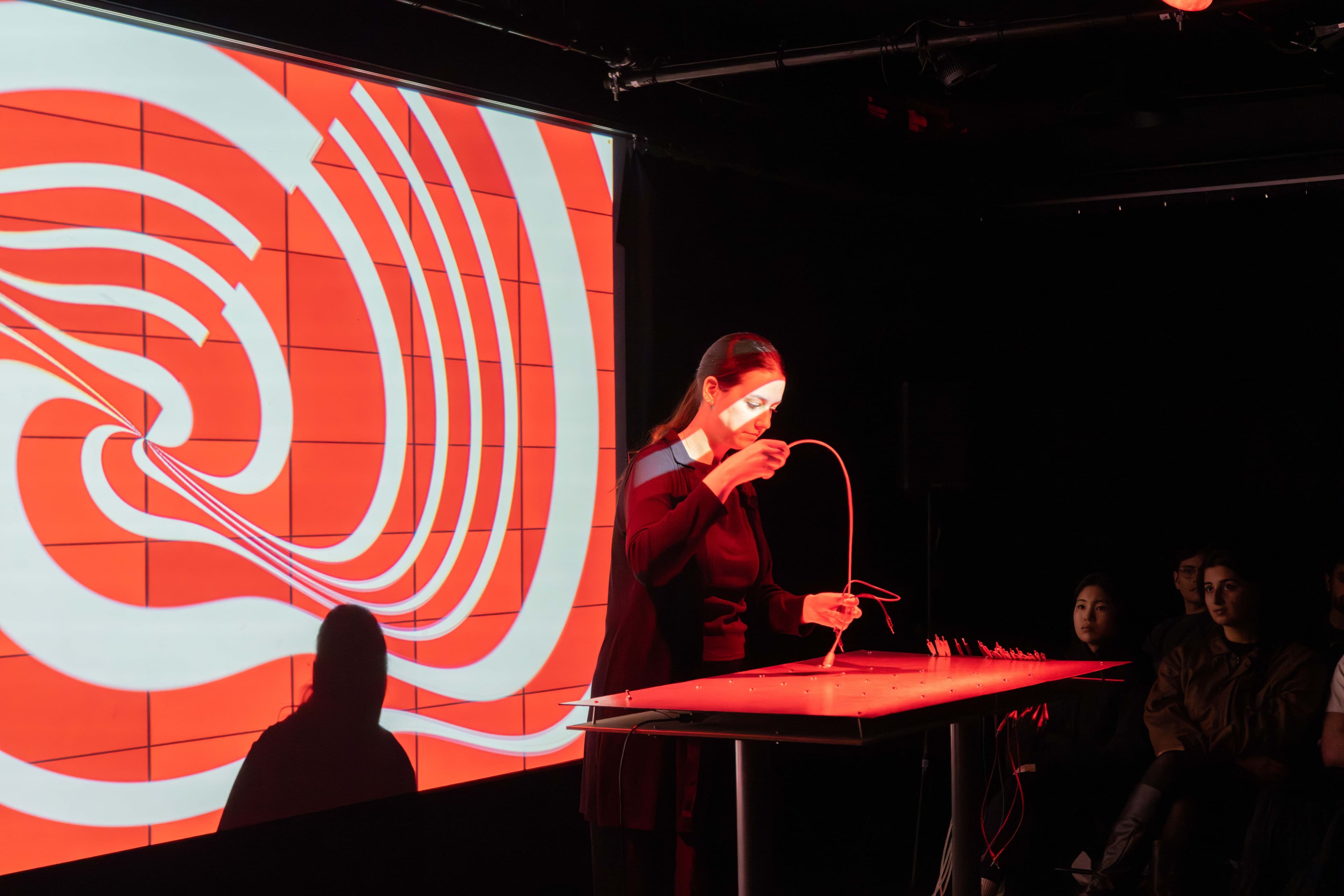 Ivana holds a cord above a table. Behind her on the screen is a projection of a red and white swirl.