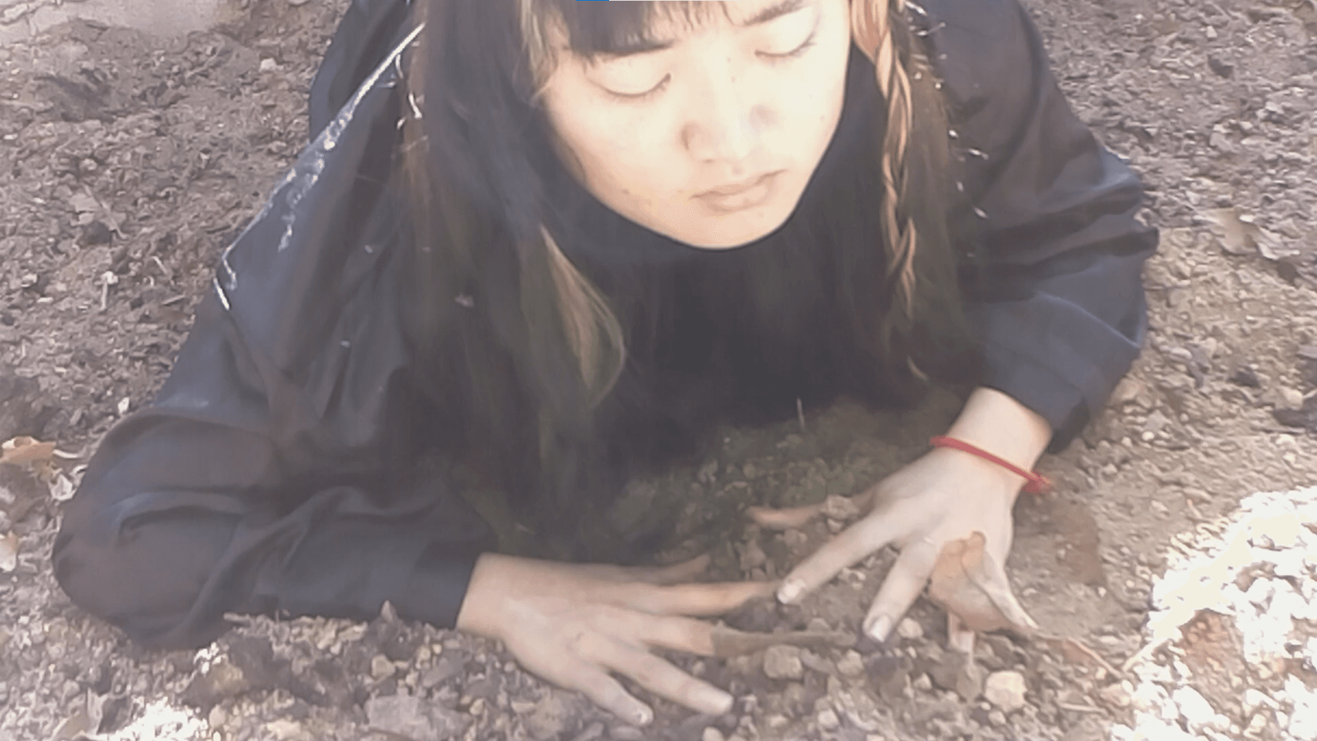 The artist lies with her eyes closed in the garden with her hands splayed out in the dirt.