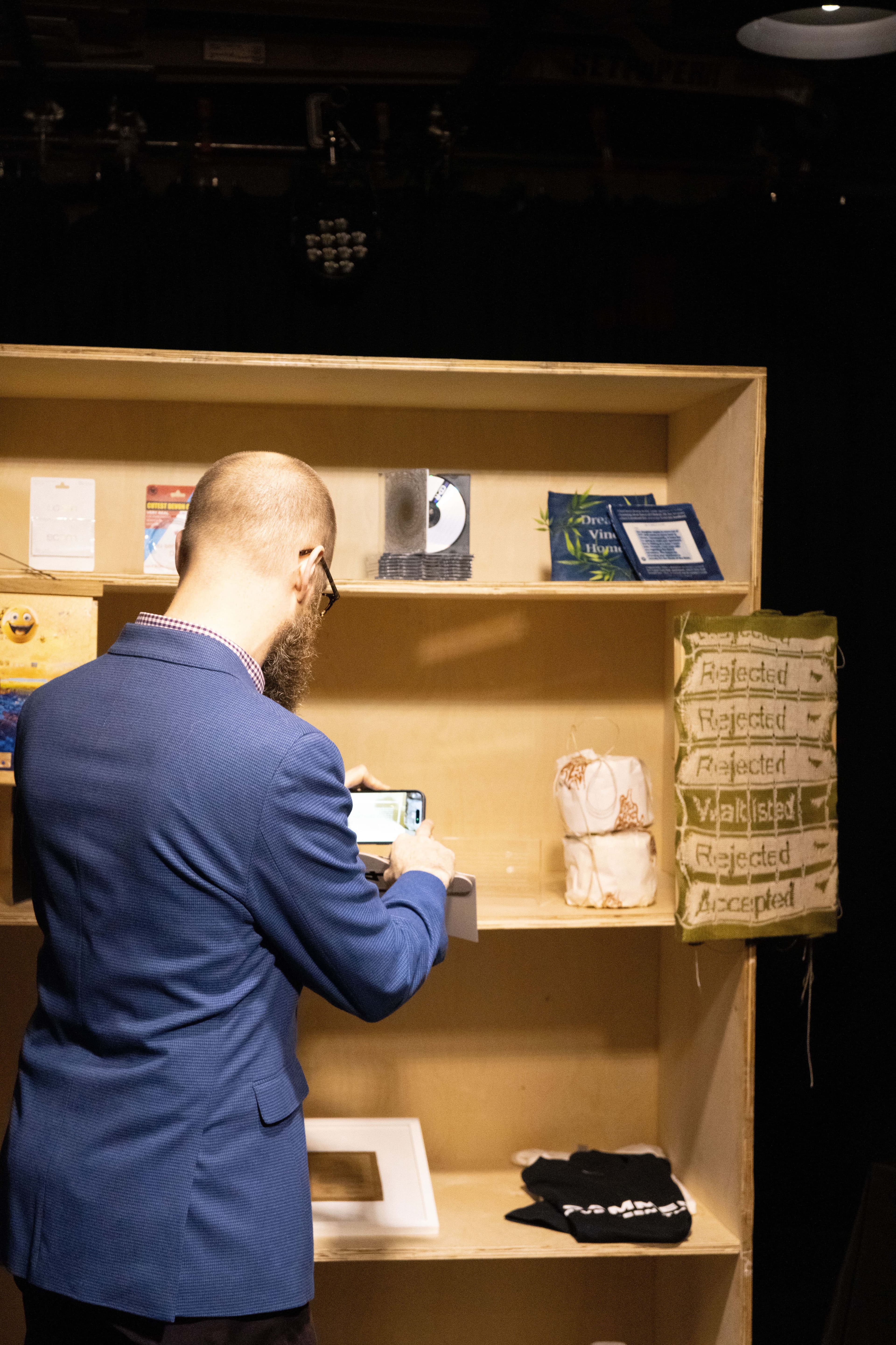 A man in a blue blazer with his back to the camera takes a picture of one of the auction items, which are arranged on a plywood bookshelf.
