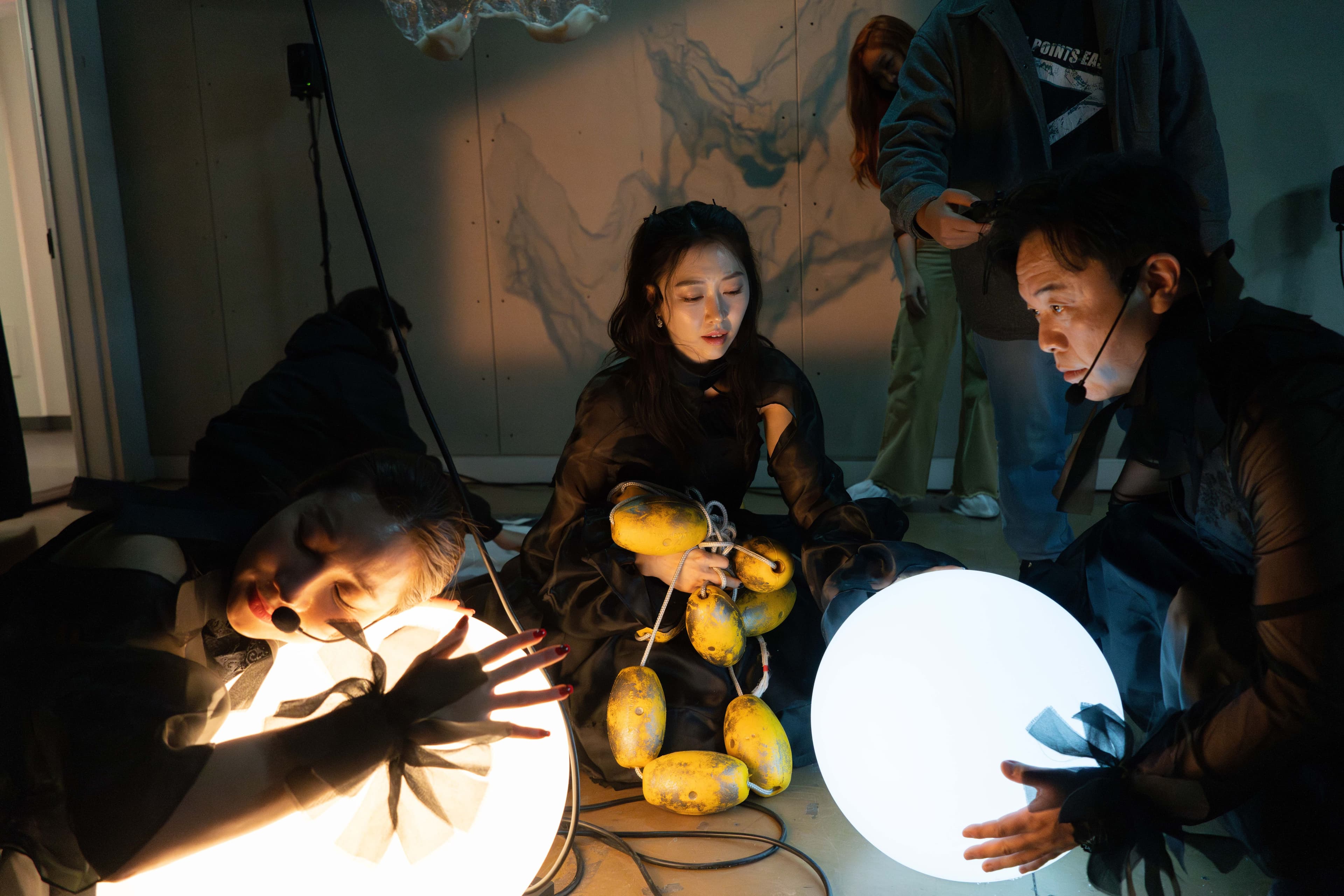 A woman presses the side of her face to a warm colored illuminated orb. Across from her a man holds another cool colored illuminated orb. Between then a woman holds a yellow buoy line.