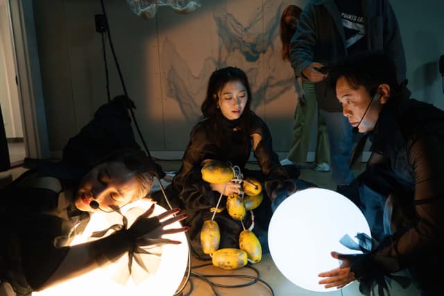 A woman presses the side of her face to a warm colored illuminated orb. Across from her a man holds another cool colored illuminated orb. Between then a woman holds a yellow buoy line.