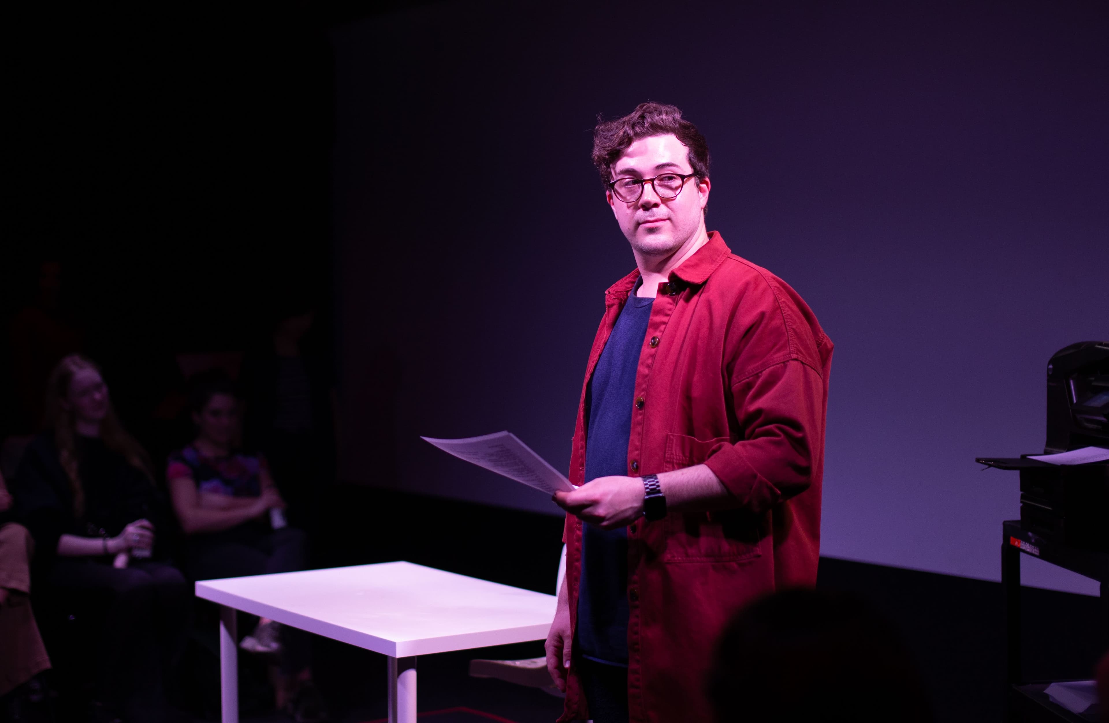 A white man with glasses wearing a red shirt holding a sheet of paper, spotlit and standing in front of a white desk.