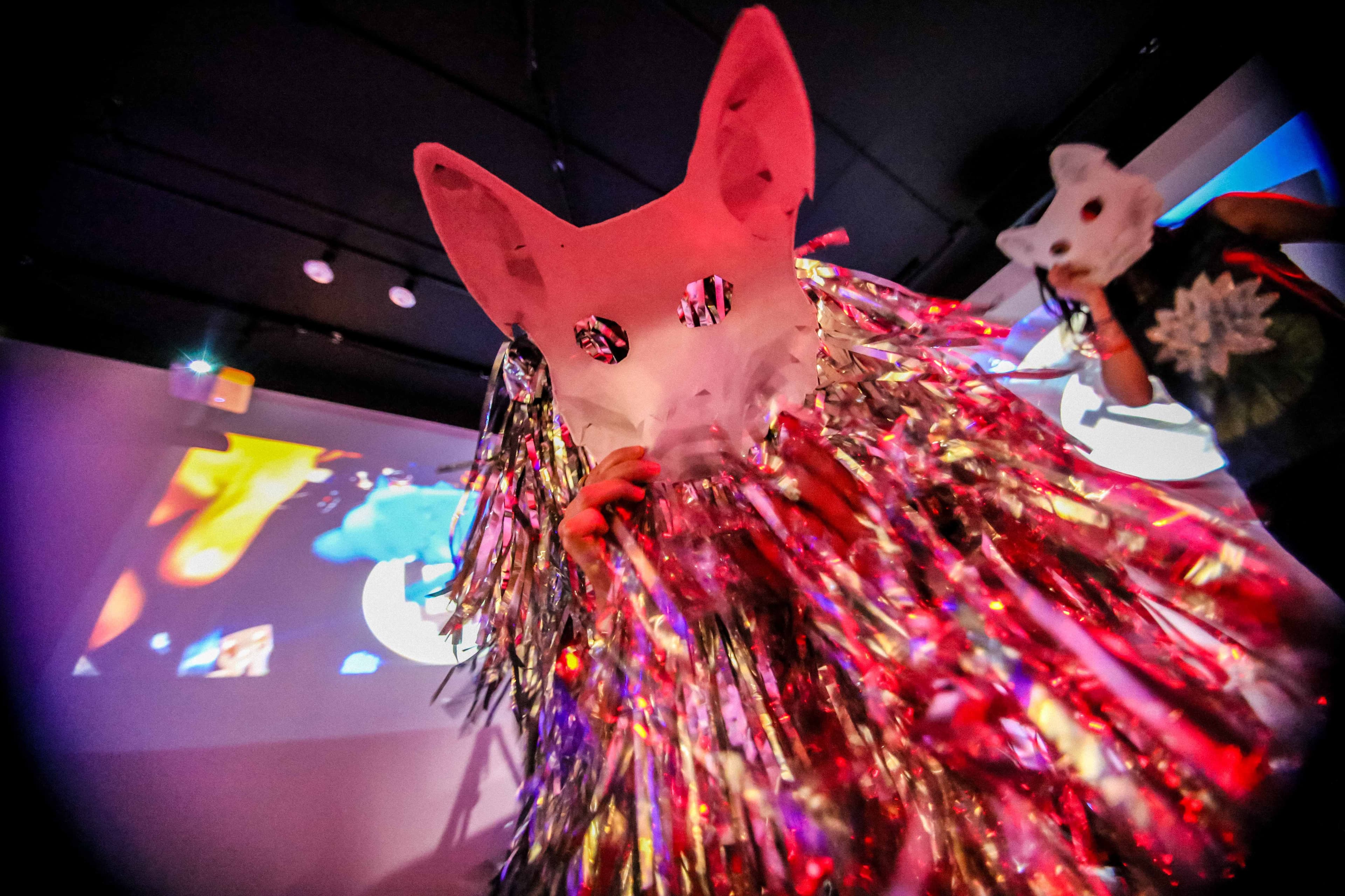 Two people wearing identical white dog masks, the person in the foreground is wearing metallic streamers that reflects the colorful lighting.