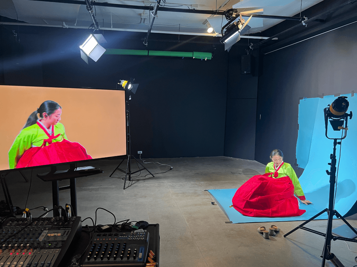 The artist Andrea Kim sits in front of a blue screen in a green and red dress, simultaneously projected on a large monitor in front of her.