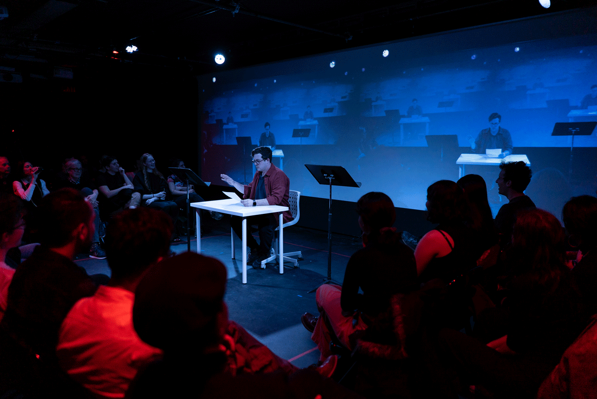 A man in a red shirt sits at a white table. He is bathed in blue light and his repeated image is project behind him. He is surrounded by a crowd bathed in red light.
