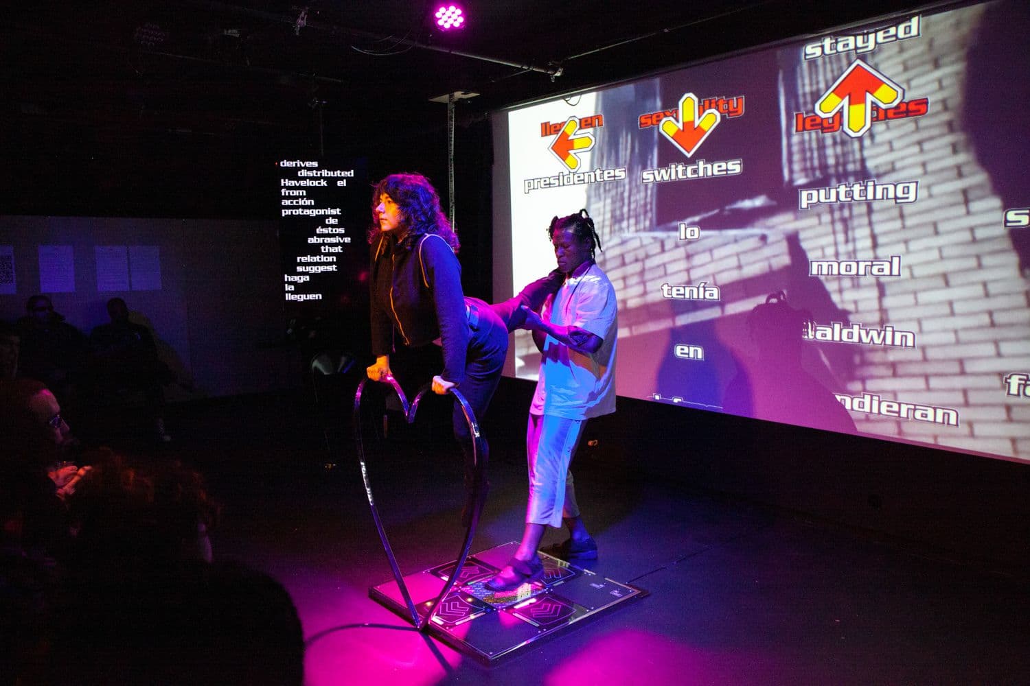 Melanie is suspended in midair and grips a metal heart. Ime holds Melanie's outstretched leg and presses their foot on the arrow of a DDR dance pad.
