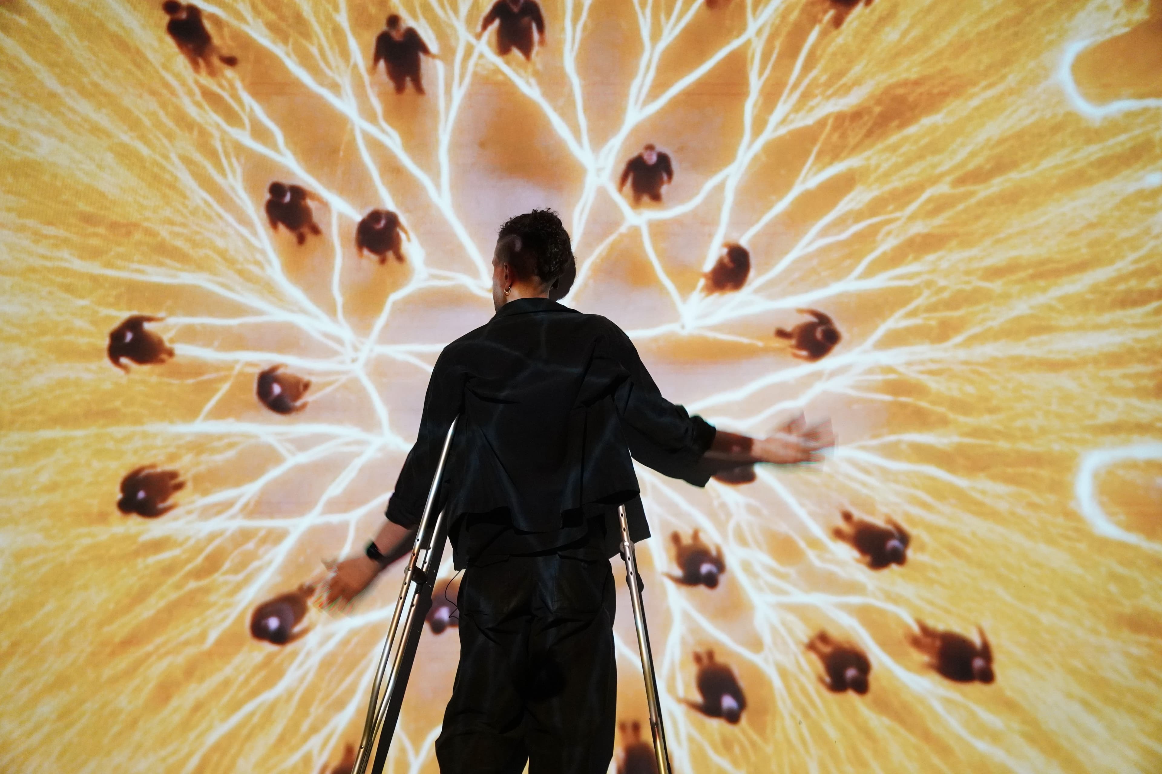 Halim stands in front of a screen holding his arms out with his back towards the camera. On screen is a yellow fractal like pattern. Tiny Halims are placed along the tendrils of the pattern