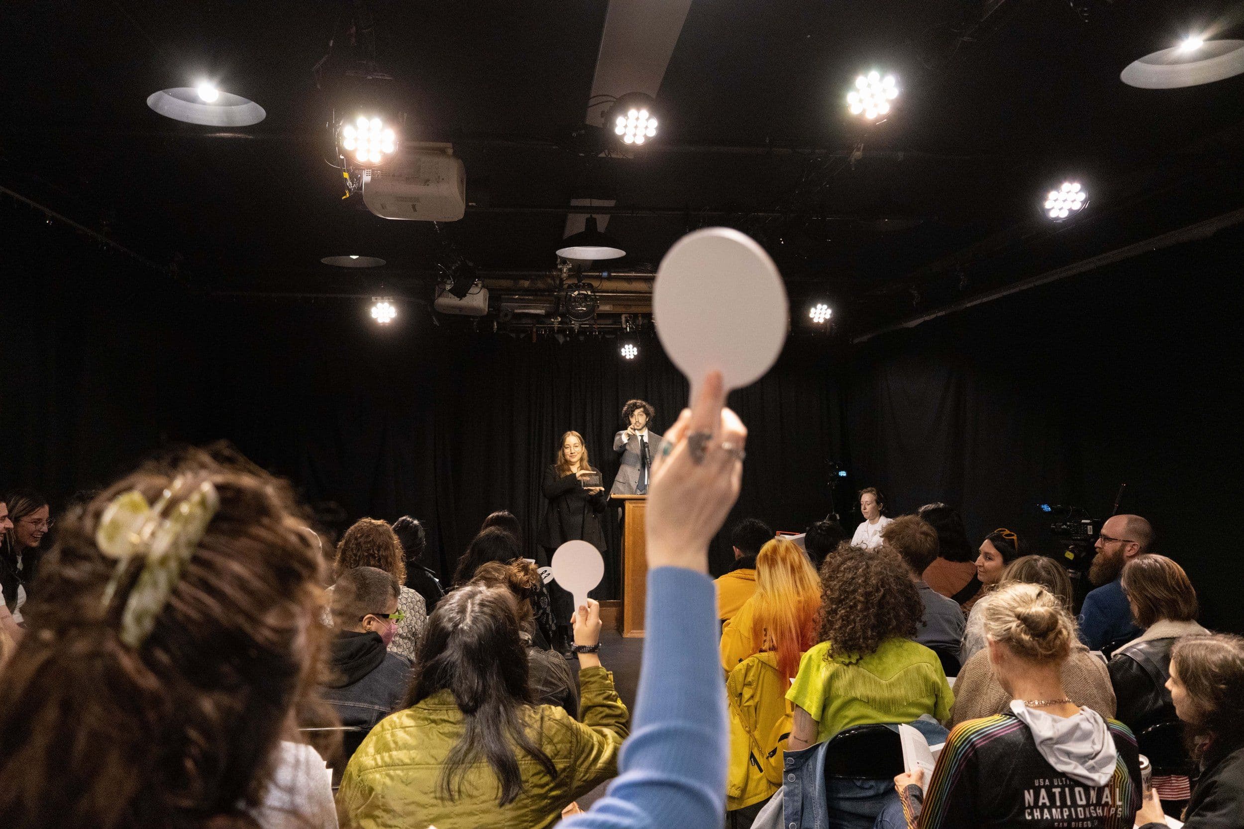 A woman wearing a light blue shirt holds up a white auction paddle. At the podium, the auctioneer, wearing a grey suit points at her, standing next to the artist Blair Simmons holding up an item for auction.
