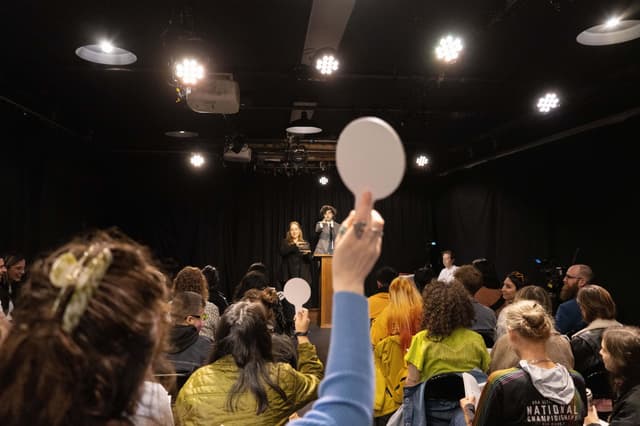 A woman wearing a light blue shirt holds up a white auction paddle. At the podium, the auctioneer, wearing a grey suit points at her, standing next to the artist Blair Simmons holding up an item for auction.