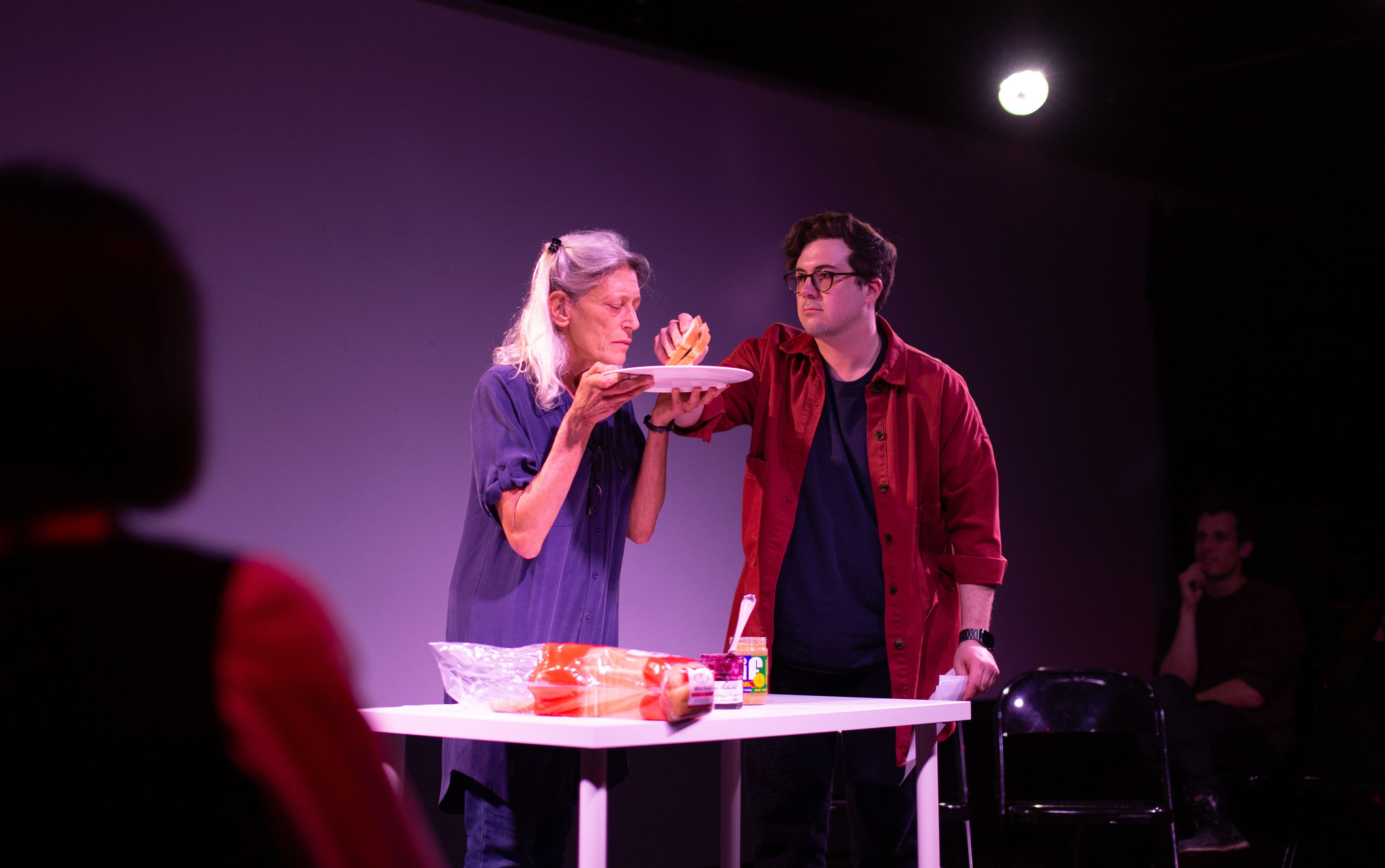 A woman in a blue jumpsuit holds a white plate up to her face. A man in a red shirt grabs a sandwich from the plate. Below on the white table there is sandwich bread, jelly and peanut butter.