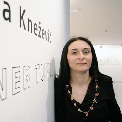 A woman with dark hair wearing a black blouse and statement necklace leans against a gallery wall with partially visible wall text that includes her last name.