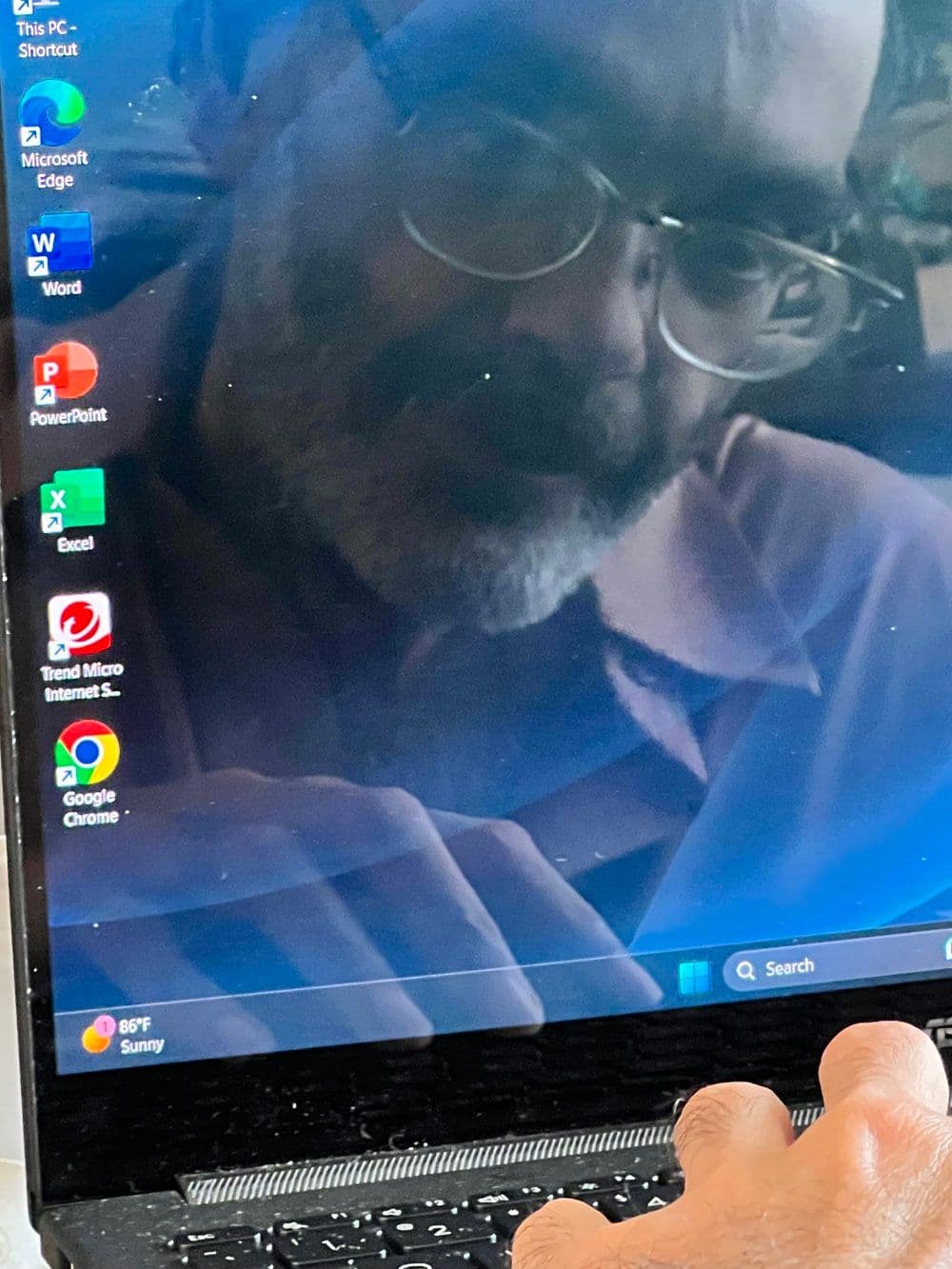 A Puerto Rican man's face wearing glasses with a short white beard is reflected on a computer desktop, with icons on the left side of the monitor. The artist's hand rests on the keyboard at the bottom of the photo.