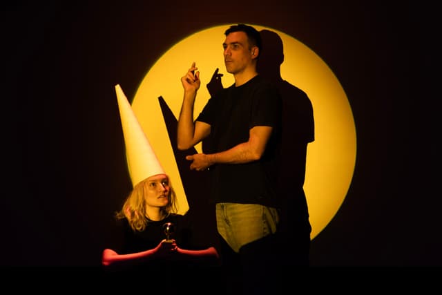 Walker Caplan kneels holding a sword, wearing a white dunce hat and looking up. Matt Romein poses holding one arm up and looking off. They are framed by a yellow spot light.