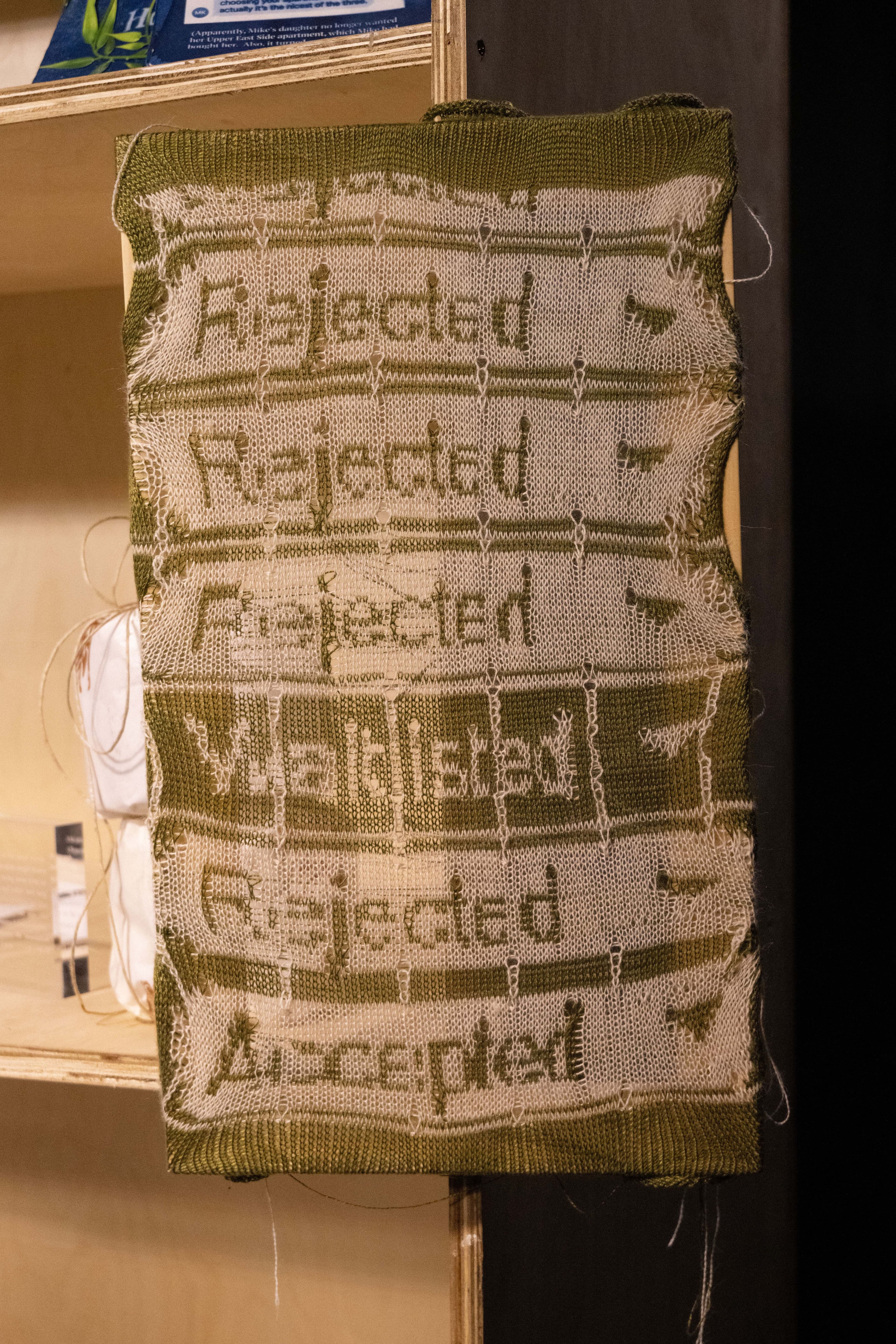 A green and white woven tapestry by e-textile artist Nicole Yi Messier, that shows a woven excerpt reading Rejected, Waitlisted, and Accepted, from a spreadsheet tracking applications to artist residencies.