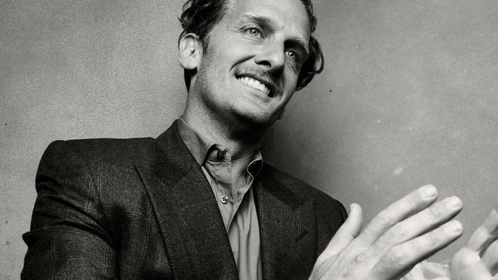 In black and white, the artist stands against a blank backdrop wearing a dark suit, his hands raised as if in applause. His head is slightly out of frame.