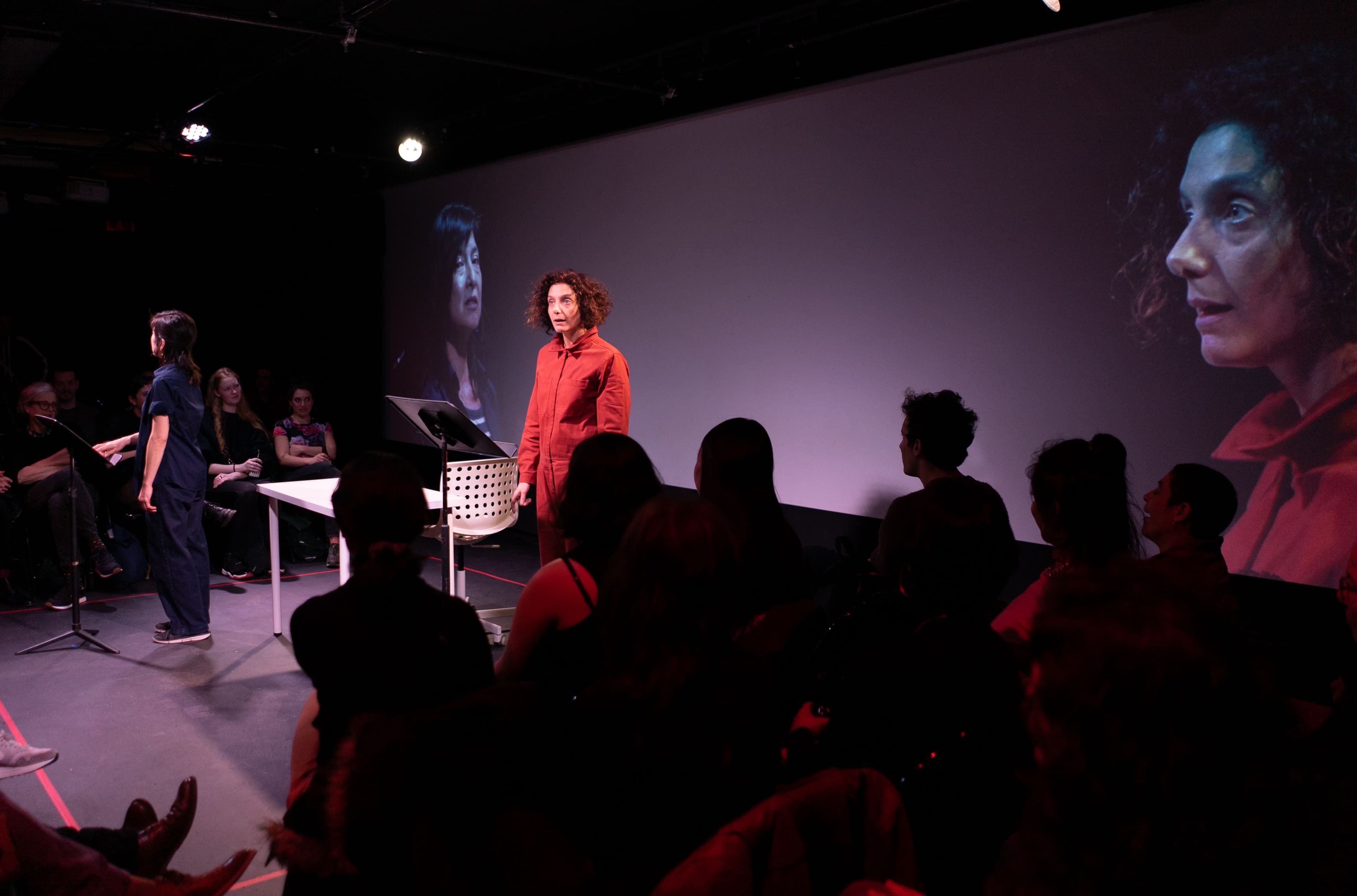 A woman in a red jumpsuit and a woman in a blue jumpsuit face outwards looking at a crowd. Their faces are projected on a wall behind them.