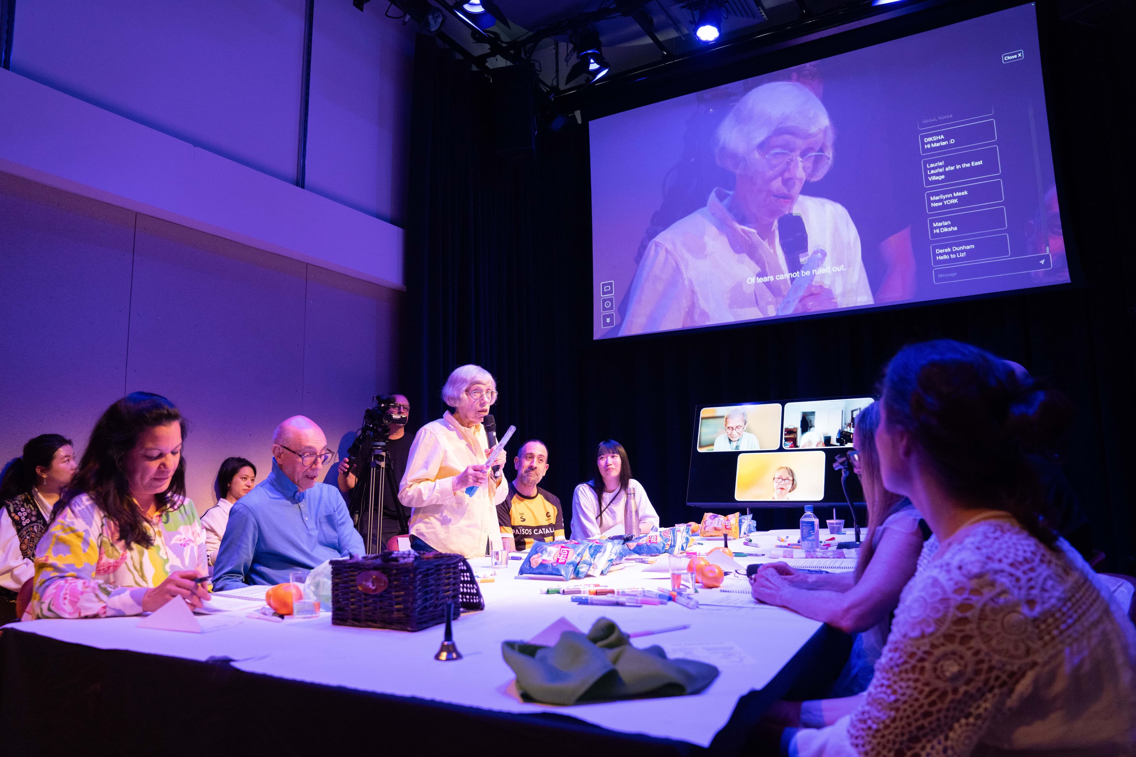 A group of older adults sit around a table together. A woman with a short white bob and glasses speaks into a microphone. Behind her on the screen is a live video feed showing the woman speaking, with the CultureHub Broadcaster interface overlayed.