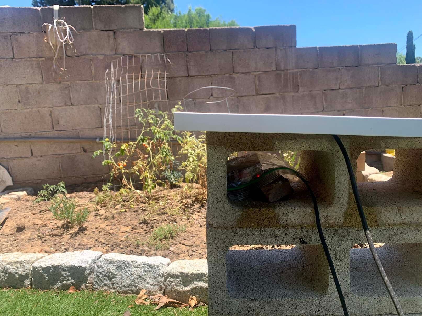 Two cables are connected to a networked camera and Raspberry Pi sitting inside a cinderblock. In the background, there is a small garden in front of a cinderblock wall.